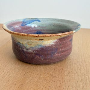May include: A handcrafted ceramic bowl with a wide rim and a rounded body. The bowl features a colour palette of burgundy, blue, and tan. The interior has a light blue and burgundy design, while the exterior is primarily burgundy.