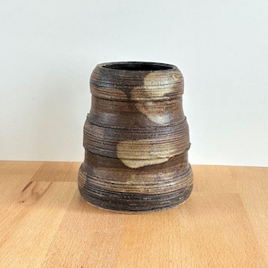 May include: A handmade ceramic vase with a cylindrical form. The vase has horizontal, textured bands in shades of brown and tan, with a dark interior. The vase sits on a wooden surface.