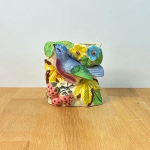 Op de afbeelding: Een keramieken plantenbak met een driedimensionale blauwe vogel op een tak. Het ontwerp bevat gebeeldhouwde groene bladeren, gele en blauwe bloemen en trossen roze aardbeien met een textuur op een crèmekleurige achtergrond.
