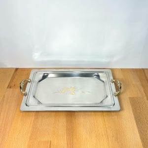 May include: A rectangular silver-toned metal serving tray with a polished finish. The tray features two ornate gold-toned metal handles on the sides and a central etched design of a cluster of fruit with leaves in the middle of the flat surface.