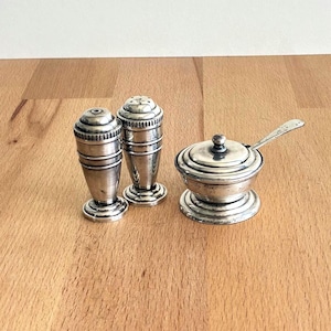 May include: An antique silver salt and pepper set with a matching lidded salt cellar and spoon. The shakers are cylindrical with decorative ridges. The salt cellar has a small spoon and a round lid. The set is displayed on a wooden surface.