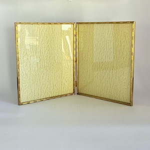 May include: A double-hinged picture frame featuring a metallic gold finish with a textured bamboo-style border. The two rectangular frames are connected by vertical hinges and contain light cream-coloured inserts with a fine, crackled texture.