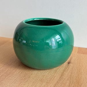 Puede incluir: Un jarrón de cerámica verde esmeralda brillante con forma redondeada y esférica. El jarrón tiene una abertura ancha y una superficie lisa y reflectante. El diseño es simple y moderno, adecuado para exhibir flores o como acento decorativo. Aproximadamente 15 cm de diámetro.