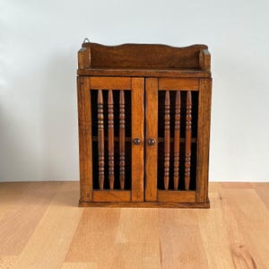 May include: A small, vintage wooden cabinet with two doors. The cabinet is made of dark brown wood and features decorative vertical spindles on the doors. It has a small shelf on top and a metal hook for hanging.