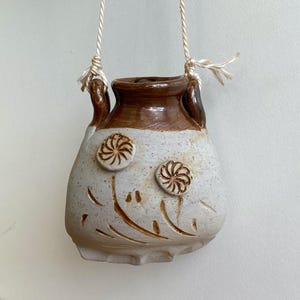 May include: A handmade, hanging ceramic planter with a cream-colored body and brown rim. The planter features two raised floral designs and etched lines. It is suspended by a natural fiber rope.