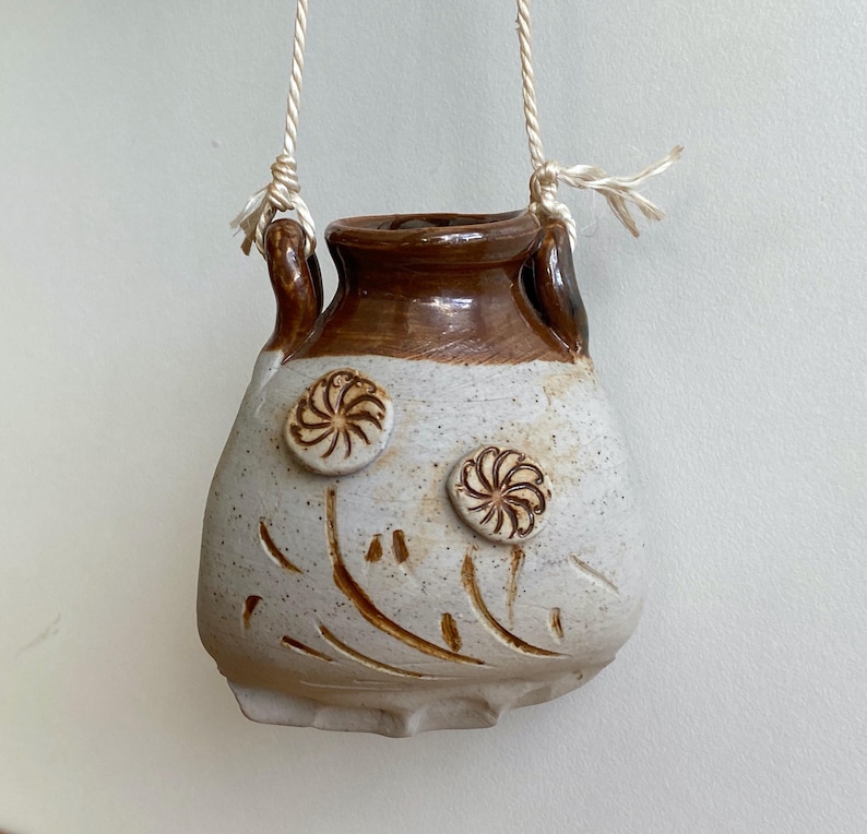 May include: A handmade, hanging ceramic planter with a cream-colored body and brown rim. The planter features two raised floral designs and etched lines. It is suspended by a natural fiber rope.