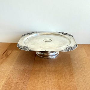 May include: A silver-coloured, square cake stand with ornate floral detailing on the corners. The stand has a circular base and a slightly raised, flat surface for displaying baked goods. The stand is made of metal and has a reflective surface.
