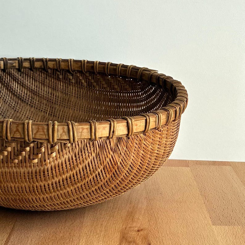 Pu&ograve; includere: Un cesto rotondo e intrecciato di colore marrone naturale. Il cesto ha un design ampio e aperto con un bordo robusto. Il motivo dell'intreccio &egrave; visibile, creando una superficie strutturata. Il cesto &egrave; posto su una superficie di legno.