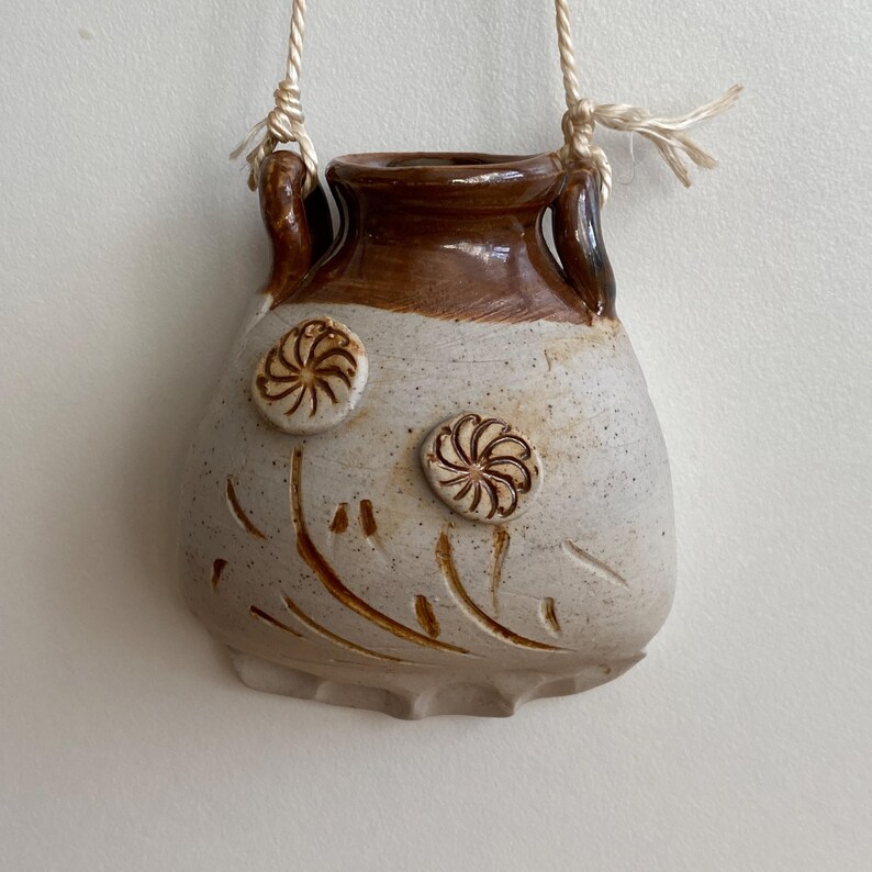 May include: A small, handmade ceramic vase with a cream-colored body and brown rim. The vase features two raised floral designs and carved leaf patterns. It hangs from a natural fiber cord.