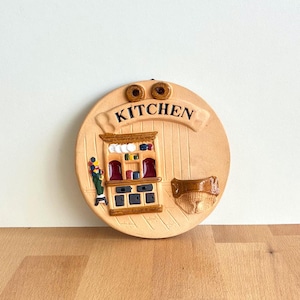 Puede incluir: Placa de pared de cocina redonda de cerámica beige. La palabra "KITCHEN" se muestra en una pancarta. La placa presenta una escena de cocina en miniatura con un armario, estantes y una cesta. La placa está decorada con detalles marrones, rojos y amarillos.