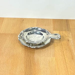 May include: A polished silver tea strainer with a matching drip tray. The strainer features a circular bowl with a perforated mesh centre and a short, curved handle. The edges of both the strainer and the tray have a scalloped, decorative rim.