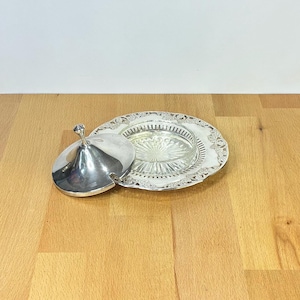 Op de afbeelding: Een verzilverde botervloot bestaande uit een rond, versierd dienblad met een decoratieve geschulpte rand en een helder glazen inzetstuk met een radiaal patroon. Een bijpassend verzilverd koepelvormig deksel met knop ligt ernaast.