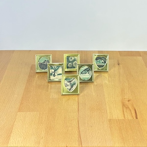Può includere: Una collezione di sei piccole cornici in metallo color oro, ognuna contenente un'illustrazione in miniatura unica. Le cornici variano nella forma, includendo aperture quadrate, ovali e a cuore, che mostrano dettagliate opere d'arte a tema naturale.