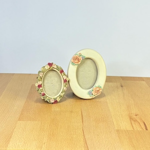 May include: Two oval-shaped picture frames in cream and off-white. The smaller frame features a dense border of sculpted pink, red, and white roses with green leaves. The larger frame has two clusters of peach roses and small blue flowers.