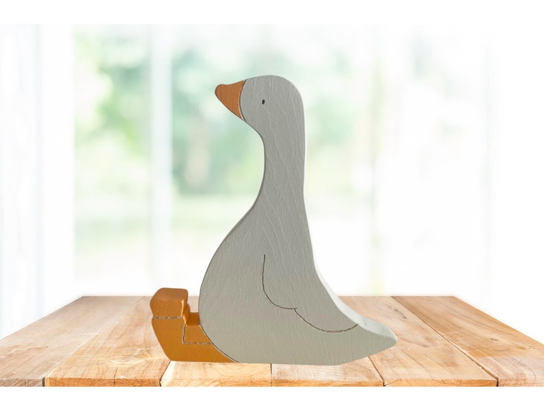 May include: A gray wooden goose figurine with an orange beak and feet. The goose is sitting with its feet tucked under its body.