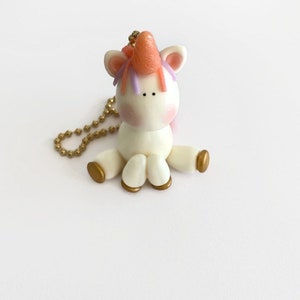May include: A handmade, seated unicorn figurine with a gold chain. The unicorn is white with a pink blush, a peach horn, and purple and pink hair. The hooves are gold. The chain is also gold.