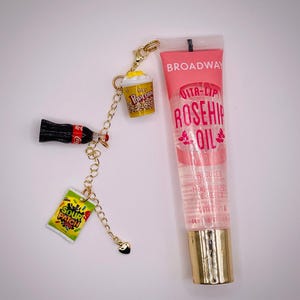 May include: A clear lip gloss tube with a gold cap and a charm featuring a popcorn bucket, a Coca-Cola bottle, and a Sour Patch Kids candy bag. The lip gloss is labeled "Broadway Vita-Lip Rosehip Oil".