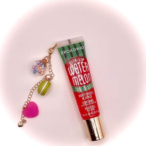 May include: A tube of Broadway Vita-Lip Watermelon Tint Gloss with a gold cap. A gold chain is attached to the tube, with a clear ball filled with colorful beads, a green and white macaron, and a pink heart charm.