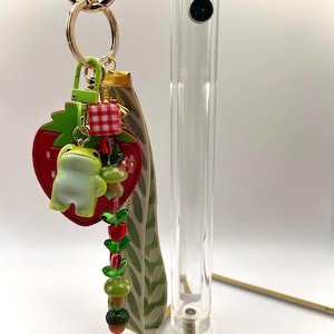 May include: A quirky keychain with a red strawberry charm, a green frog figurine, and a beaded strand with mushroom and leaf details. The keychain has a gold-tone chain and a green clasp.