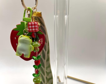 May include: A whimsical keychain featuring a red strawberry charm, a green frog figurine, and a beaded strand with mushroom and leaf accents. The keychain includes a gold-tone chain and a green clasp.