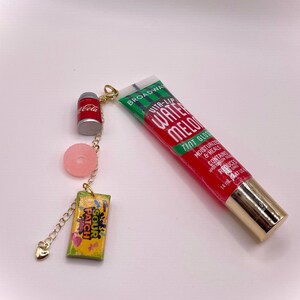 May include: A gold chain featuring charms: a red Coca-Cola can, a pink sweet, and a Sour Patch Kids candy packet. A red and green tube of Broadway Vita-Lip Watermelon Tint Gloss is also present.