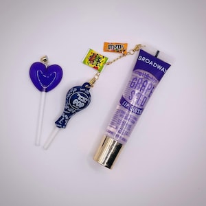May include: An assortment of items including a purple heart-shaped lollipop, a Tootsie Roll Pop charm, a tube of Broadway Grape Seed Lip Gloss, and candy. The lip gloss tube has a gold cap and the text "GRAPE SEED LIP GLOSS".