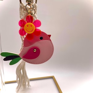 May include: A pink bird-shaped keychain with a red and yellow flower charm. The bird has a red beak and a dark pink wing. The keychain is attached to a macrame cord and a gold-colored clasp.