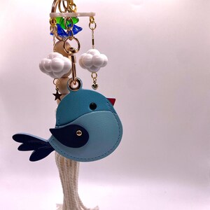 May include: A light blue bird-shaped keychain with a dark blue wing and accents. The keychain includes white cloud charms, a gold star, and a gold-colored clasp. The bird has a red beak and a small gold eye.