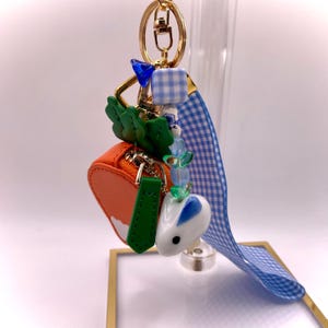 May include: A gold-colored keychain with a variety of charms. The charms include a small orange purse, a white bunny with a blue heart, green leaves, and blue and white checkered beads. A blue ribbon is attached.