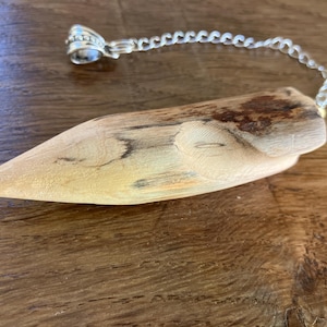 Op de afbeelding: Een houten pendel met een zilverkleurige ketting en een decoratieve bovenkant. De pendel is gemaakt van lichtgekleurd hout met donkerbruine markeringen. De ketting is bevestigd aan een zilverkleurige ring.