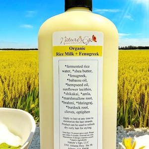 May include: A white plastic bottle with a black cap, labeled "Nature's Ego Organic Rice Milk + Fenugreek". The bottle is in front of a field of yellow flowers. The label lists the ingredients, which include fermented rice water, shea butter, fenugreek, babassu oil, hempseed oil, sunflower lecithin, shikakai, amla, marshmallow root, brahmi, bhringraj, burdock root, cloves, and optiphen. The bottle is 32 ounces (947 ml).