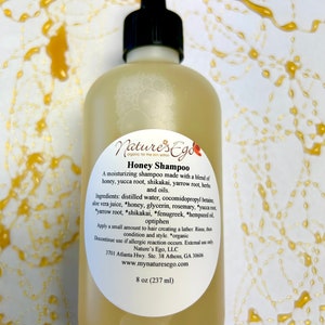 May include: A clear amber glass bottle of honey shampoo with a black cap and a white label. The label reads "Nature's Ego" and "Honey Shampoo". The bottle is sitting on a white surface with a yellow liquid splatter.
