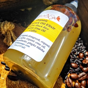 May include: A clear glass bottle with a yellow label that reads "Organic Greek Oil Elixir, Organic Black Rice, Organic Emu Oil, Coffee". The bottle is filled with a light yellow liquid and is sitting on a wooden surface with black and brown coffee beans.