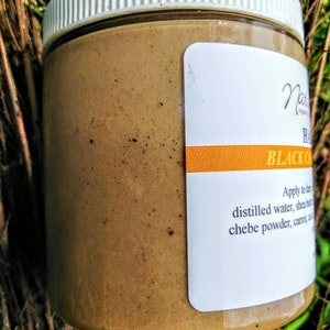May include: A glass jar filled with a brown, thick paste. The jar has a white lid and a label that reads "Black Castor Oil Hair Mask" and "Apply to damp hair with distilled water, shea butter, chebe powder, carrot oil, and honey."