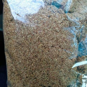 May include: A close-up of a brown, granular substance in a clear plastic bag. The substance appears to be a type of spice or seasoning.