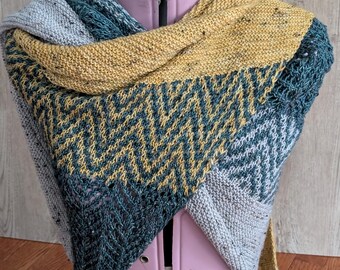 Puede incluir: Un chal hecho a mano, multicolor, con un patrón de chevron. El chal presenta tonos de gris, verde azulado y amarillo mostaza. El accesorio de punto se muestra en un maniquí rosa, mostrando su diseño y textura.