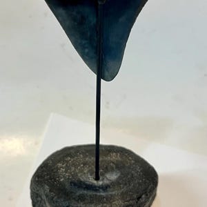 Pu&ograve; includere: Un dente di squalo fossilizzato grigio scuro montato su una sottile asta di metallo nero, inserita in una base cilindrica di vertebra fossilizzata grigio scuro. Un righello di legno &egrave; visibile in basso, mostrando che misura oltre 15 cm di altezza.