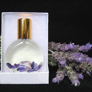 May include: A clear glass bottle with a gold lid filled with a clear liquid and small purple and pink stones. The bottle is in a white box with a white background. A bunch of lavender flowers are on the right side of the image.