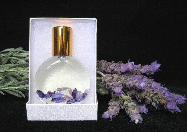 May include: A clear glass bottle with a gold lid filled with a clear liquid and small purple and pink stones. The bottle is in a white box with a white background. A bunch of lavender flowers are on the right side of the image.