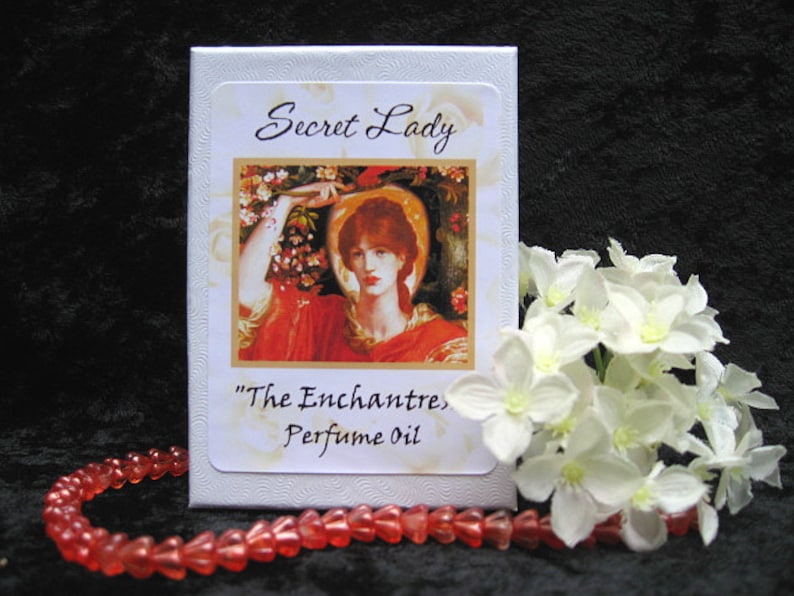 Może przedstawiać: Białe pudełko z kwiatowym wzorem i tekstem "Secret Lady" i "The Enchantress Perfume Oil".
