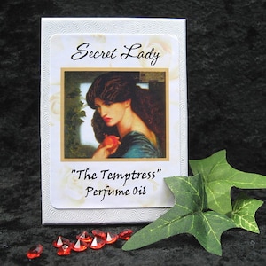 May include: White box with a white label that says "Secret Lady" and "The Temptress" Perfume Oil. The label has a picture of a woman with dark hair and a red apple in her hand.