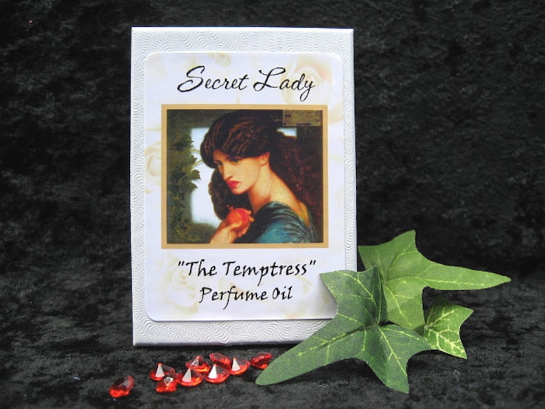 May include: White box with a white label that says "Secret Lady" and "The Temptress" Perfume Oil. The label has a picture of a woman with dark hair and a red apple in her hand.