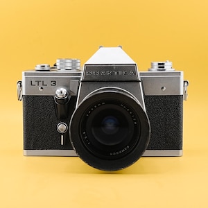 May include: A vintage Praktica LTL 3 camera with a black lens and silver body. The camera has the word "PRAKTICA" printed on the top and "LTL 3" on the side. The lens has the text "CAMERON" printed on it.