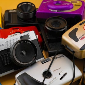 Puede incluir: Una colección de cámaras vintage coloridas, incluyendo una cámara plateada y negra con una lente de 50 mm, una cámara roja con el texto "MX-35" y una cámara púrpura. Las cámaras están dispuestas sobre una superficie amarilla.