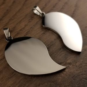 Puede incluir: Dos colgantes de plata con forma de mitades de coraz&oacute;n. Los colgantes son lisos y brillantes, y tienen un dise&ntilde;o simple y elegante.