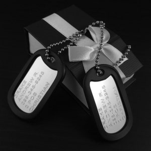 May include: Two black dog tags with silver lettering. The tags are engraved with the text "Drake Andrew R. 088-345-8245 POS CHRISTIAN". The tags are connected by a silver chain and are sitting on top of a black box with a white ribbon.