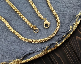 May include: Two gold chains with a braided design, lying on a gray stone surface.