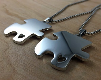 May include: Two silver puzzle piece pendants with heart cutouts on a silver chain. The pendants are connected together.