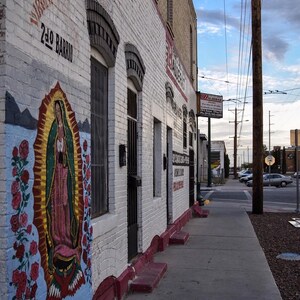 May include: Exterior view of a white brick building with a mural of the Virgin Mary and roses. The building has arched windows and a sidewalk in front. The sky is cloudy.