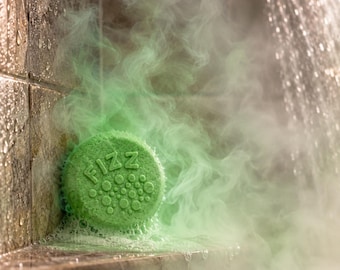 May include: A round, green shower steamer tablet with the word FIZZ embossed on the front, surrounded by a cluster of small circles. The tablet is actively dissolving, releasing a thick, green-tinted steam into the air while resting on a ledge.